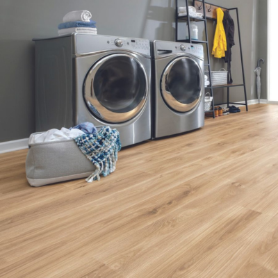 A modern laundry room with light oak wood-look flooring, featuring silver appliances, a black storage rack, and a grey laundry basket.